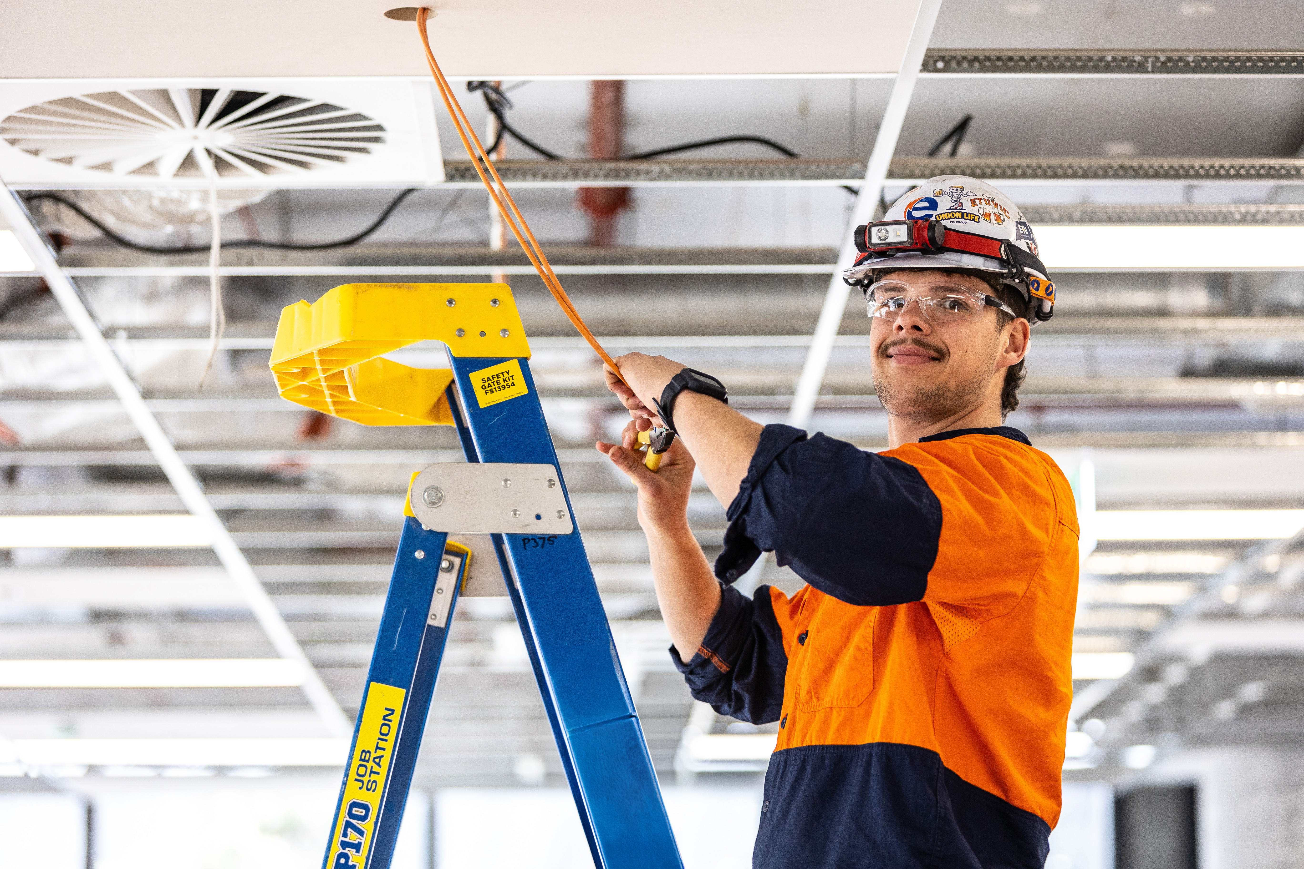 electrical fit out safety glasses electrician electrical work wiring young man working on electrical work indoors commercial building site construction site blue-collar worker building clothing engineer engineering hard hat ladder man person candidates labour hire high vi vest