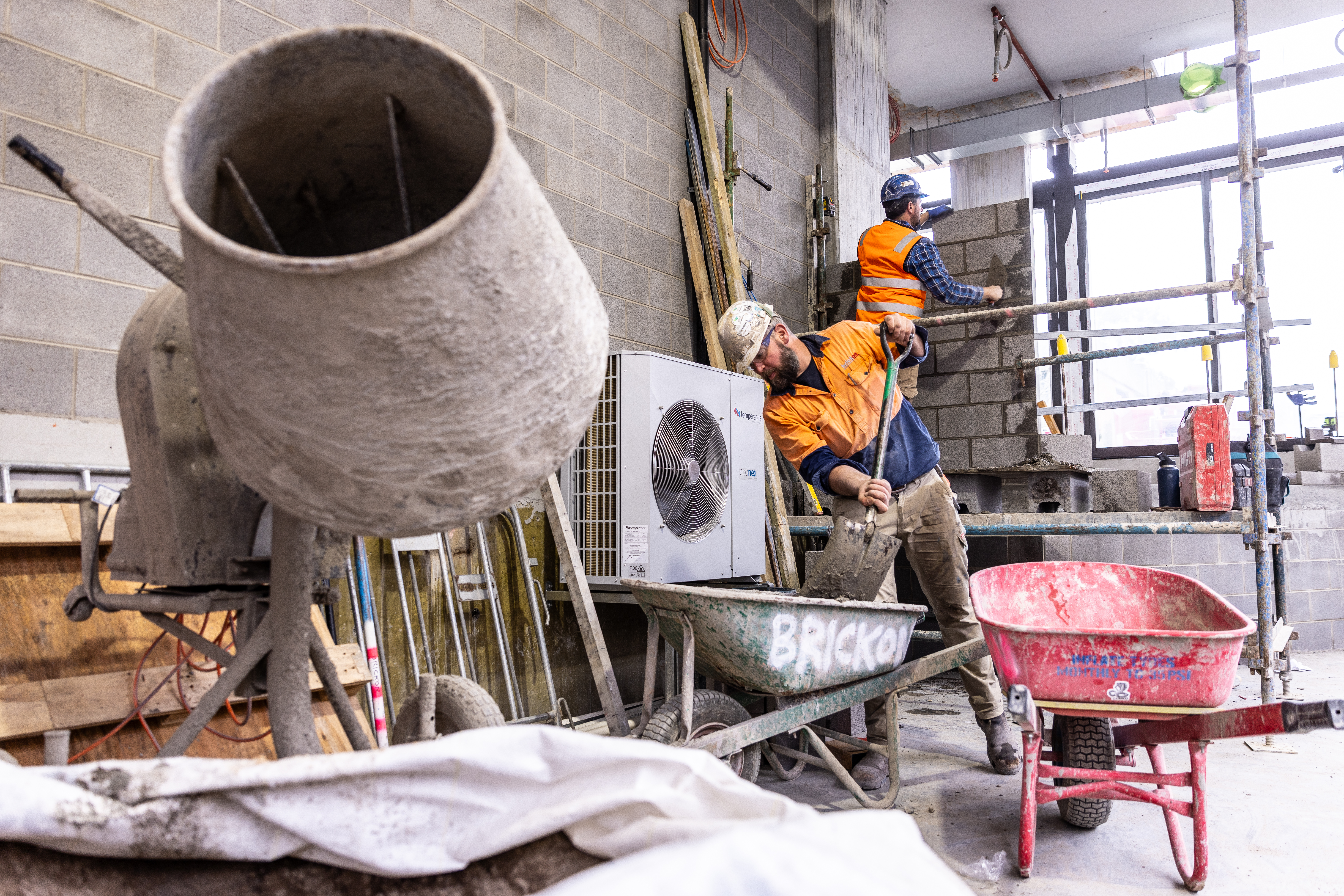 bricklaying brick layer mortar wheelbarrows two men mixing mortar and laying bricks on construction site indoors scaffolding concrete mixer indoor pipe transport employers construction worker labour hire window high vis vest tiles hardhat